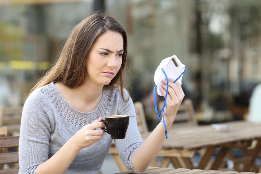 Doubtful woman looking at protective mask on a coffee shop - HospitalityLawyer.com®