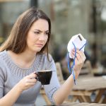 Doubtful woman looking at protective mask on a coffee shop - HospitalityLawyer.com®