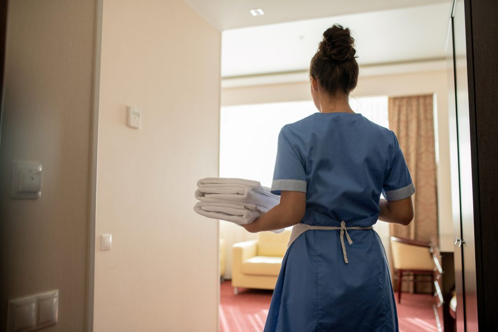 Back view of young chamber maid in uniform carrying stack of clean white towels - HospitalityLawyer.com® Back view of young brunette chamber maid in uniform carrying stack of clean white towels for guests while entering one of rooms