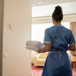 Back view of young chamber maid in uniform carrying stack of clean white towels - HospitalityLawyer.com® Back view of young brunette chamber maid in uniform carrying stack of clean white towels for guests while entering one of rooms