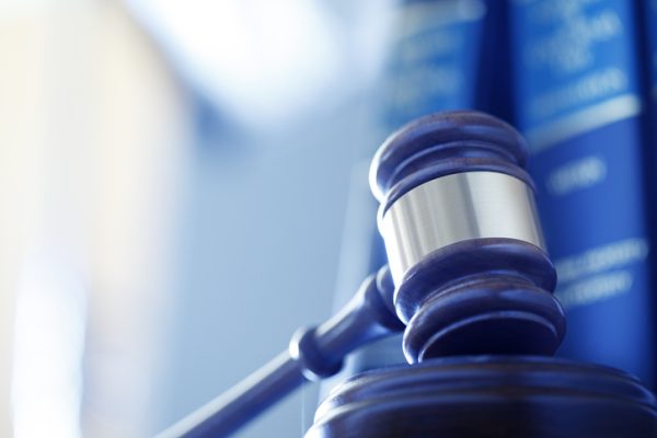 A wooden gavel rests on its sounding block in front of a row of law books and an office interior that are out of focus in the background. Photographed using a shallow depth of field.
