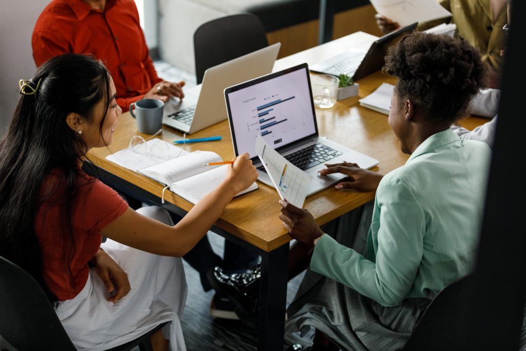 Group of business people discussing project reports during a meeting - HospitalityLawyer.com® High angle view of two young businesswomen reviewing a document and data on laptop during a meeting with their colleagues.