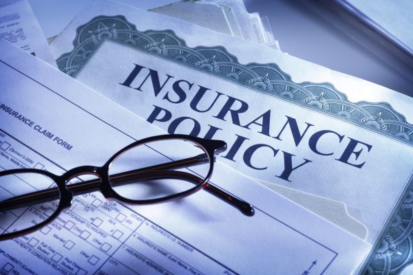 A pair of glasses on a health insurance policy claim form. This photograph is taken with a cool blue light and a shallow depth of field. The reading glasses rest on the numerous forms one must fill out for health insurance. "Insurance Policy" reads clear and big across the document.
