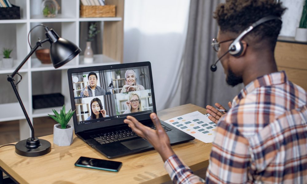 Multiethnic group of people having business meeting through video call on laptop. View from shoulder of black man sitting at home office and working remotely.