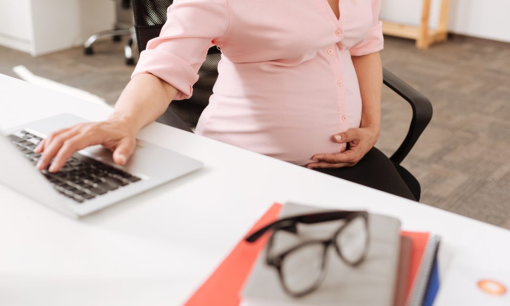 Pleasant pregnant woman sitting at the table and touching her belly while using her laptop