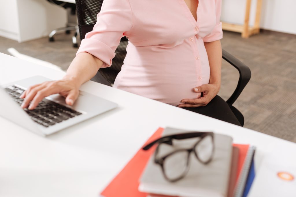 Professional pregnant office worker sitting in at the table - HospitalityLawyer.com® Pleasant pregnant woman sitting at the table and touching her belly while using her laptop