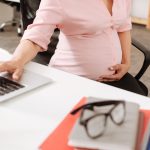 Pleasant pregnant woman sitting at the table and touching her belly while using her laptop