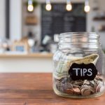 Tips jar on coffee shop table - HospitalityLawyer.com® Foreground focus is on a glass jar labeled "tips" in chalk. The jar is sitting to one side on a rustic wooden table, full of coins and bills, with coffee shop scenery in the background.