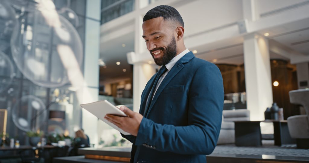 Businessman digital tablet or strategy planning in hotel conference lobby or airport travel lounge Happy smile corporate worker or technology app to budget company finance or schedule target goals - HospitalityLawyer.com® Businessman, digital tablet or strategy planning in hotel conference lobby or airport travel lounge. Happy smile, corporate worker or technology app to budget company finance or schedule target goals
