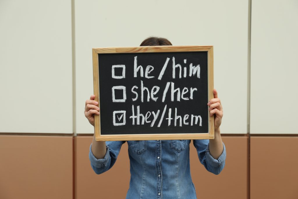 Woman holding chalkboard with list of gender pronouns near color wall - HospitalityLawyer.com® Woman holding chalkboard with list of gender pronouns near color wall