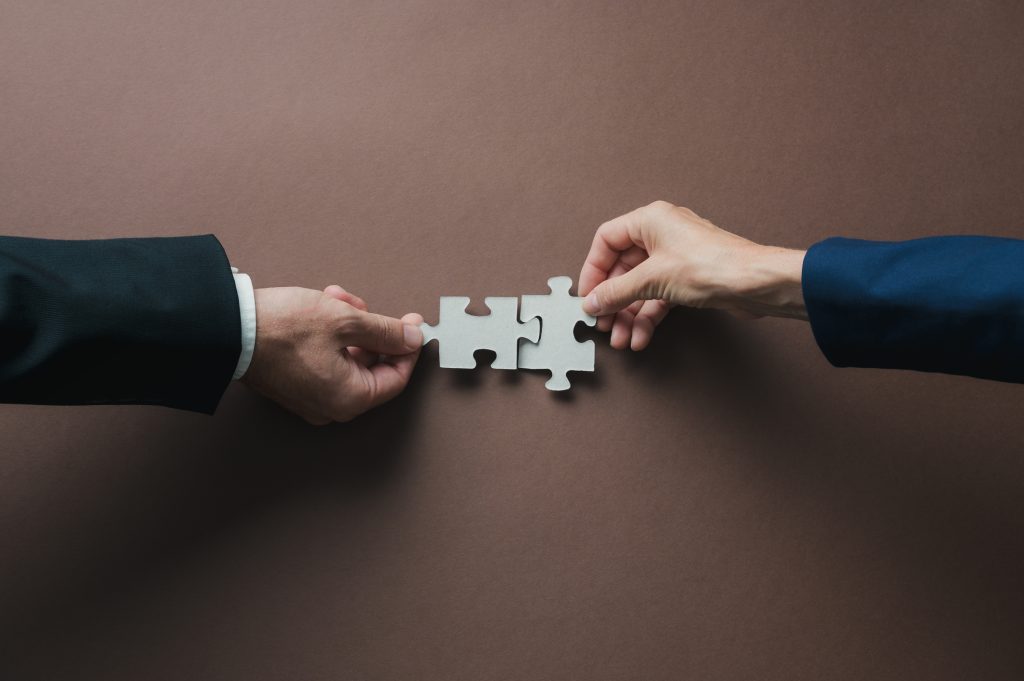 Teamwork and partnership conceptual image - HospitalityLawyer.com® Teamwork and partnership conceptual image - hands of a businessman and businesswoman joining blank matching puzzle pieces over brown background.