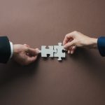 Teamwork and partnership conceptual image - HospitalityLawyer.com® Teamwork and partnership conceptual image - hands of a businessman and businesswoman joining blank matching puzzle pieces over brown background.