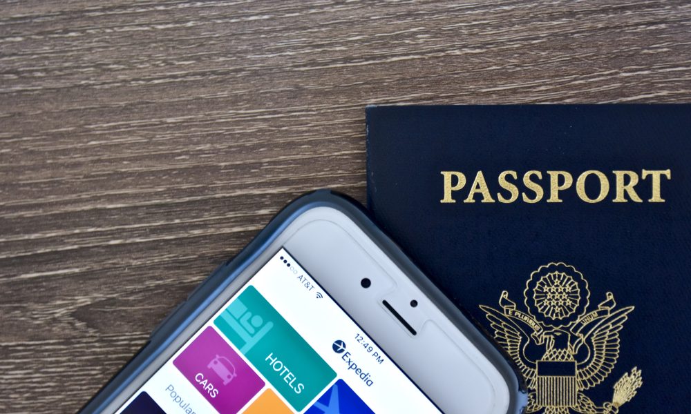 An Apple iPhone 6s displaying the Expedia application homepage on the screen while laying next to an American passport.