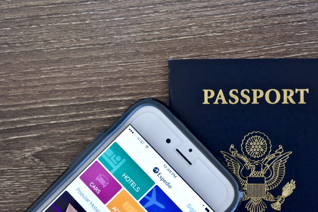 Passport and iPhone with expedia app - HospitalityLawyer.com® An Apple iPhone 6s displaying the Expedia application homepage on the screen while laying next to an American passport.
