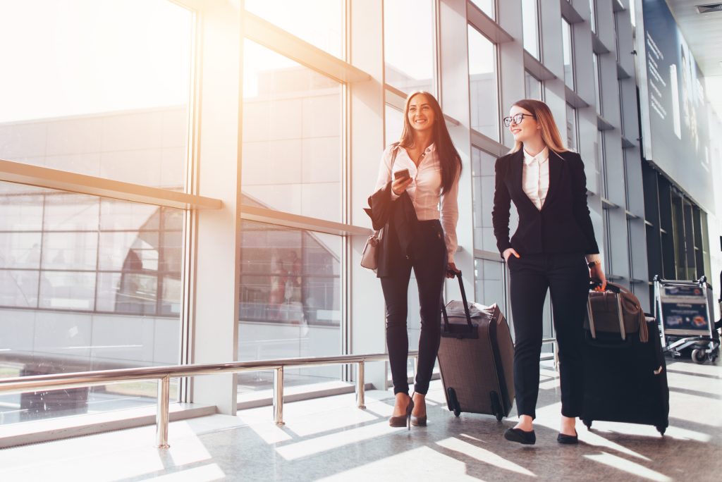 Two smiling business partners going on business trip carrying suitcases while walking through airport passageway - HospitalityLawyer.com®