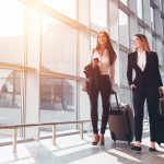 Two smiling business partners going on business trip carrying suitcases while walking through airport passageway - HospitalityLawyer.com®