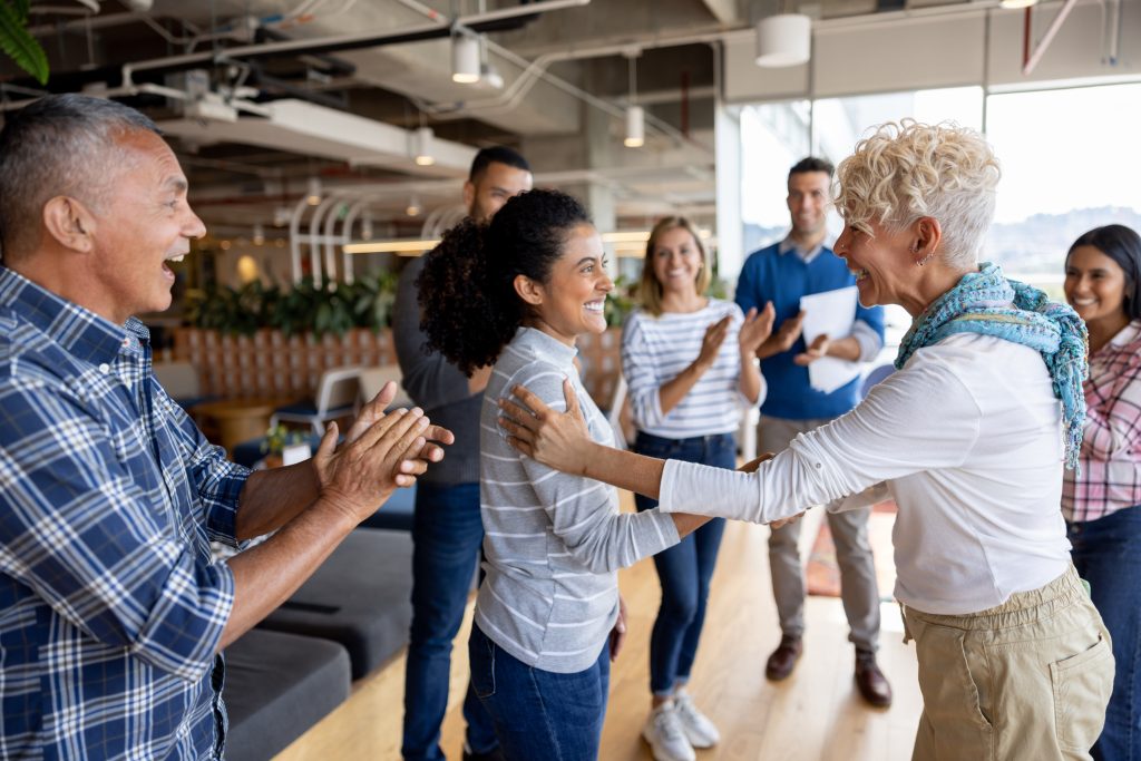 Boss giving woman a promotion at the office and team clapping for her - HospitalityLawyer.com®