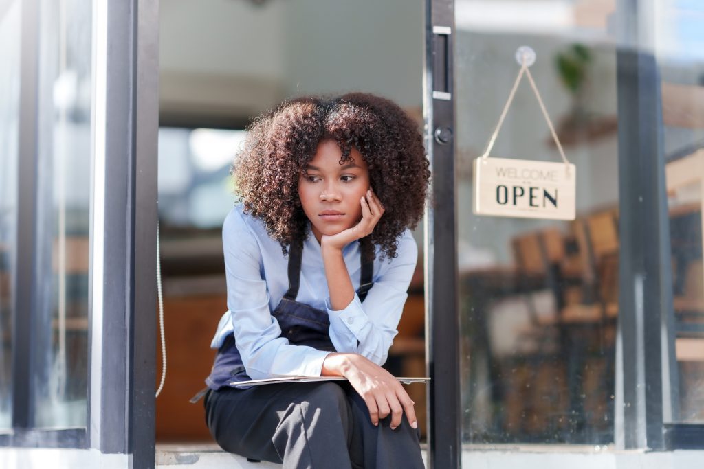 Female shopkeeper sitting stressed out at the store entrance frustrated by the economic impact - HospitalityLawyer.com® Female shopkeeper sitting stressed out at the store entrance frustrated