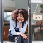 Female shopkeeper sitting stressed out at the store entrance frustrated by the economic impact - HospitalityLawyer.com® Female shopkeeper sitting stressed out at the store entrance frustrated