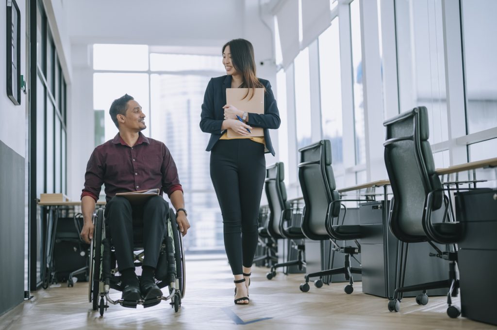 Indian white collar male worker in wheelchair having cheerful discussion conversation with his female asain chinese colleague coworking at walkway corridor - HospitalityLawyer.com®