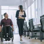 Indian white collar male worker in wheelchair having cheerful discussion conversation with his female asain chinese colleague coworking at walkway corridor - HospitalityLawyer.com®