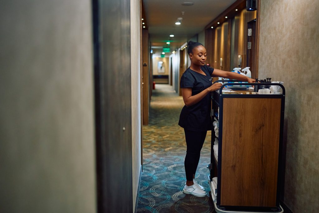 Black female housekeeper replenishing toiletries in guest rooms in a hotel - HospitalityLawyer.com® Black female housekeeper replenishing toiletries in guest rooms in a hotel