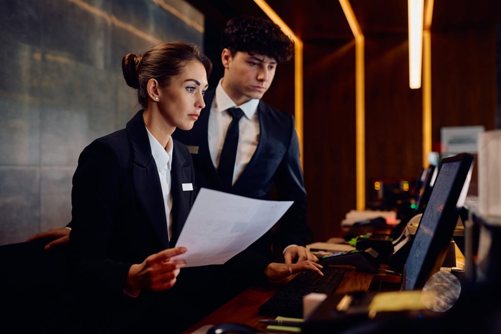 Two receptionists working on a computer while checking documents at hotel reception desk - HospitalityLawyer.com®