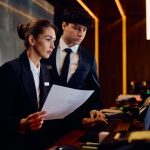 Two receptionists working on a computer while checking documents at hotel reception desk - HospitalityLawyer.com®
