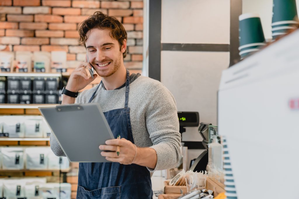 Cheerful young good-looking barista talking on phone holding folder in loft coffee shop - HospitalityLawyer.com®