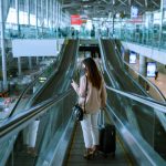 Asian businesswoman traveler with face mask on the move using escalator in airport - HospitalityLawyer.com®