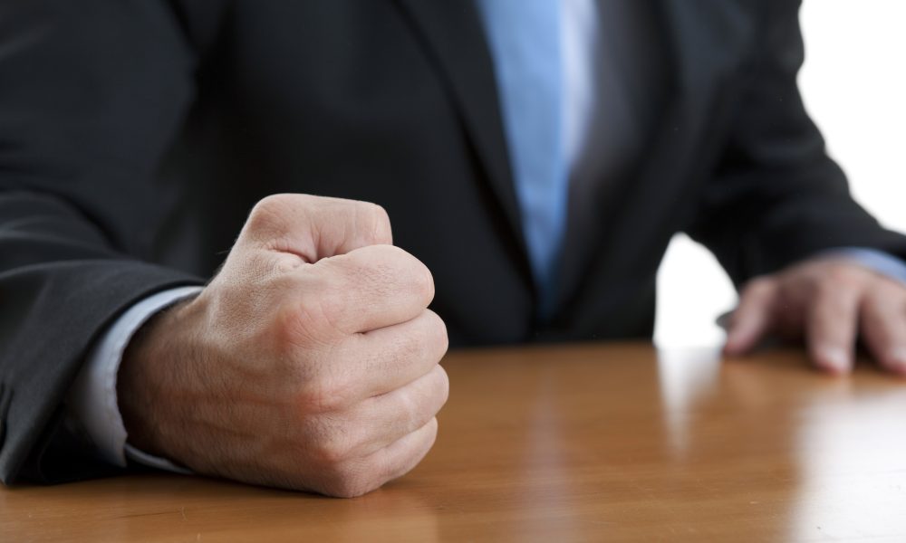 Close-up of a clenched fist resting on a wooden table, with a person in a dark suit and blue tie in the background.