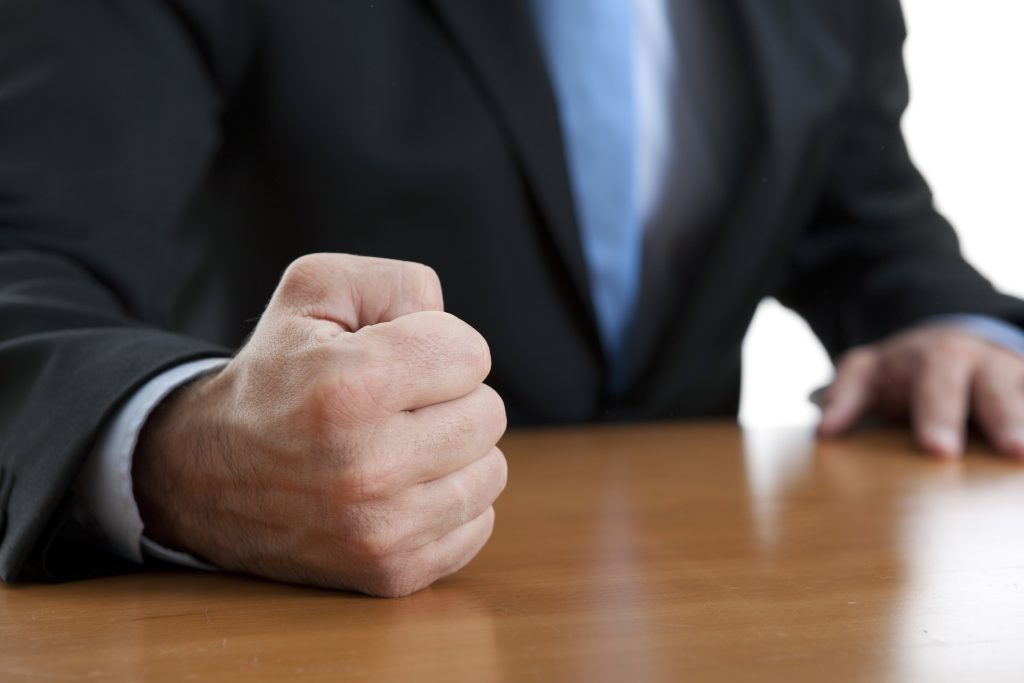 Close-up of a clenched fist resting on a wooden table, with a person in a dark suit and blue tie in the background.