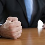 Close-up of a fist of a furious businessman on a table - HospitalityLawyer.com® Close-up of a clenched fist resting on a wooden table, with a person in a dark suit and blue tie in the background.