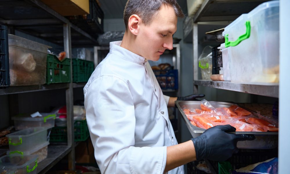 Chef in a white coat with gloves places a tray of raw salmon in a refrigerated storage shelf in a walk-in cooler.