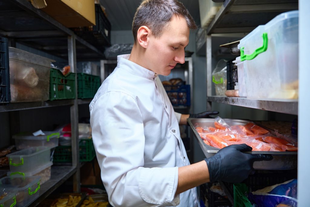 Kitchen employee with a tray in a restaurant refrigerator - HospitalityLawyer.com® Chef in a white coat with gloves places a tray of raw salmon in a refrigerated storage shelf in a walk-in cooler.