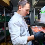 Kitchen employee with a tray in a restaurant refrigerator - HospitalityLawyer.com® Chef in a white coat with gloves places a tray of raw salmon in a refrigerated storage shelf in a walk-in cooler.
