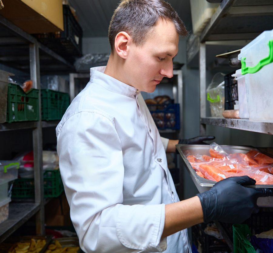 Chef in a white coat with gloves places a tray of raw salmon in a refrigerated storage shelf in a walk-in cooler.