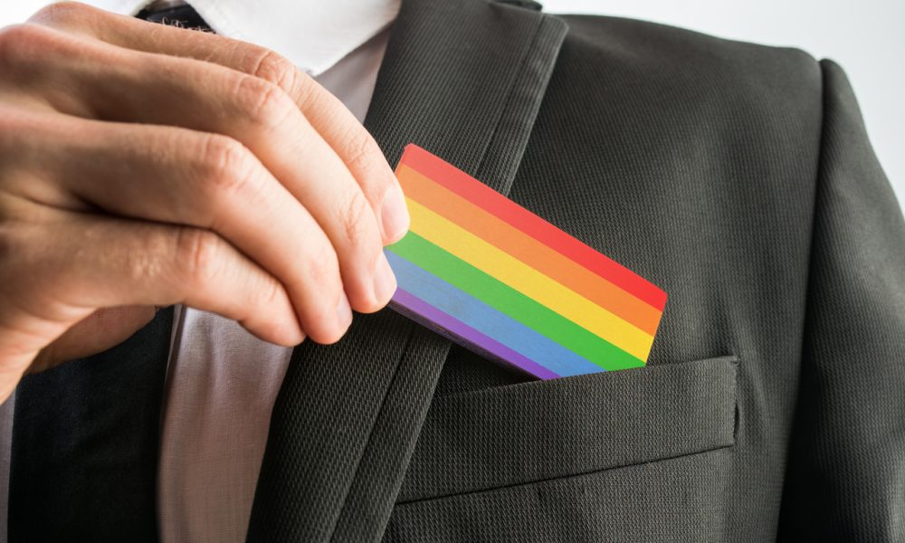Person in a suit slides a rainbow pride card into the breast pocket, signaling LGBTQ+ pride in a professional setting.