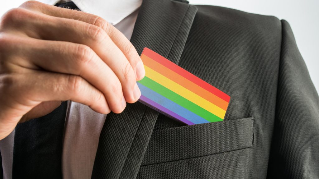 Man withdrawing wooden card painted as the gay pride flag - HospitalityLawyer.com® Person in a suit slides a rainbow pride card into the breast pocket, signaling LGBTQ+ pride in a professional setting.