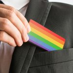 Man withdrawing wooden card painted as the gay pride flag - HospitalityLawyer.com® Person in a suit slides a rainbow pride card into the breast pocket, signaling LGBTQ+ pride in a professional setting.