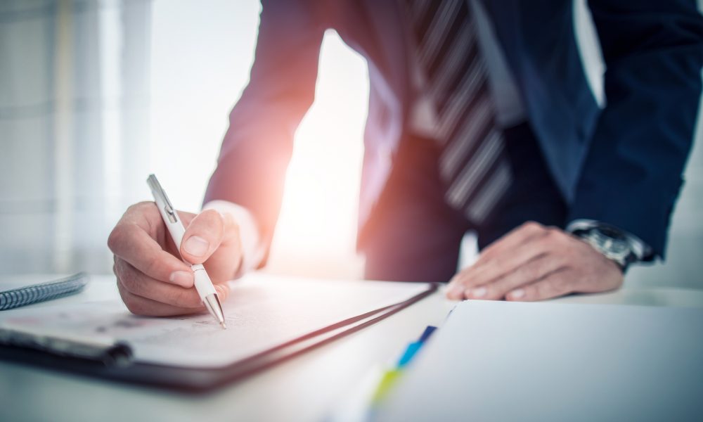 Close-up of a person in a suit signing a document with a white pen at a desk in an office setting