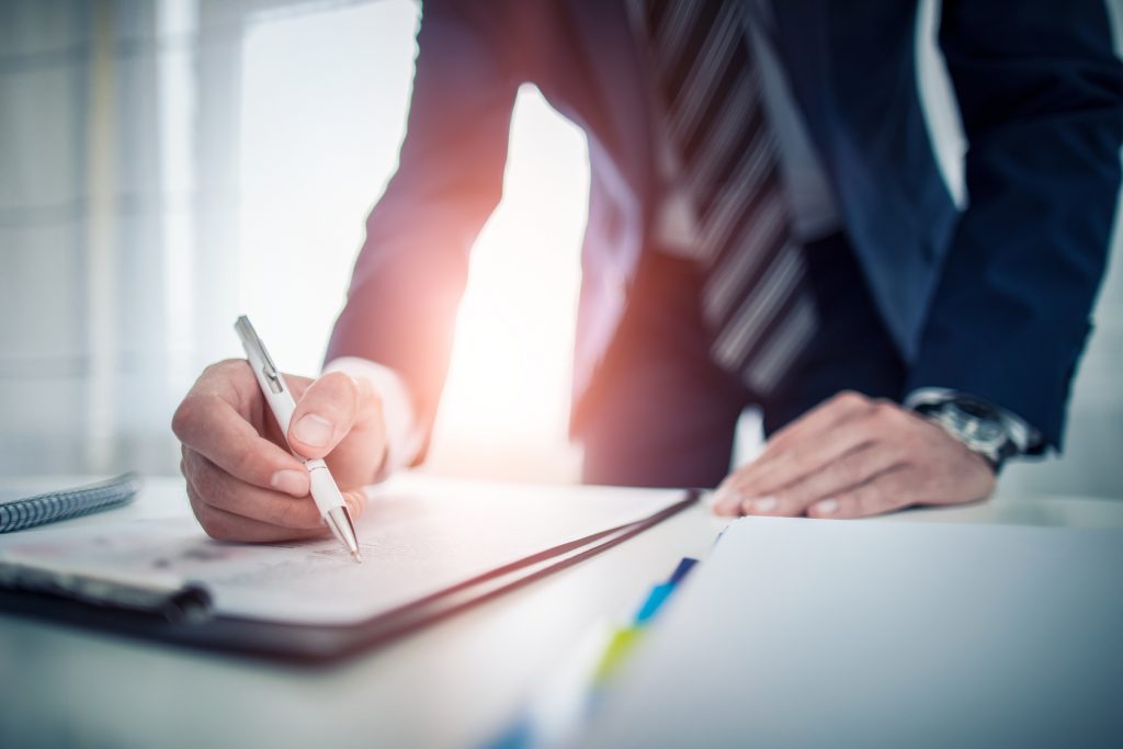 Business man signing contract making a deal - HospitalityLawyer.com® Close-up of a person in a suit signing a document with a white pen at a desk in an office setting