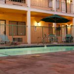 Pool and Rooms at Dusk - HospitalityLawyer.com® Outdoor motel/pool area with teal lounge chairs, a large green umbrella, and warm wall lights along a two-story building with balconies at dusk